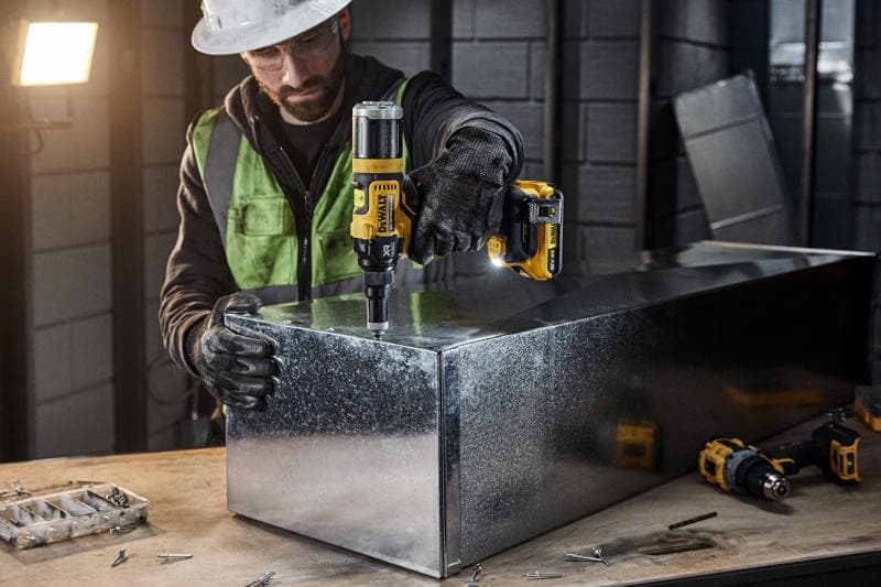 Uma pessoa com equipamento de segurança utiliza uma ferramenta elétrica sem fio DEWALT DCF403NT para trabalhar em uma caixa de metal em uma oficina industrial. Diversos parafusos e ferramentas são visíveis na bancada de trabalho.