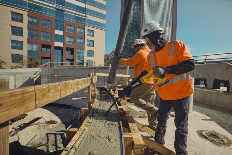Dois operários da construção vestindo capacetes de segurança e camisetas laranja de alta visibilidade estão trabalhando em uma obra. Um usa uma ferramenta vibradora de concreto DEWALT no concreto recém despejado, enquanto o outro direciona uma mangueira de concreto. Prédios modernos de escritórios aparecem ao fundo.