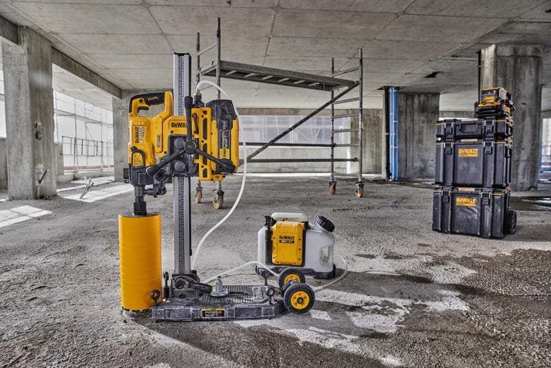 Uma máquina de perfuração de núcleo DEWALT com um acessório de broca cilíndrico amarelo e um tanque de água conectado está posicionada no chão de um canteiro de obras de concreto. Ao fundo, há andaimes e caixas empilháveis da DEWALT.