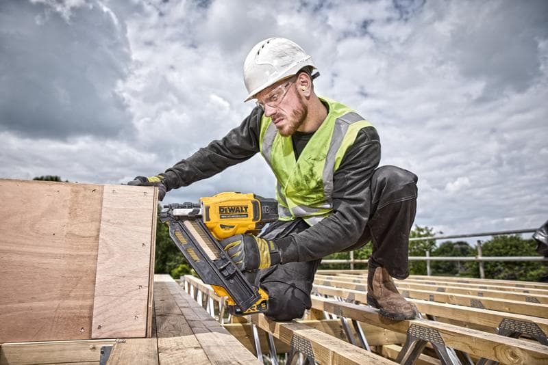 Um trabalhador da construção civil usando capacete de segurança e colete de alta visibilidade está utilizando uma pistola de pregos amarela DEWALT DCN930N para fixar tábuas de madeira em um canteiro de obras ao ar livre. O rosto do trabalhador está desfocado para proteger a privacidade.