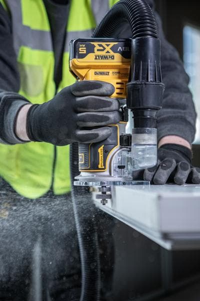 Uma pessoa usando luvas de segurança e colete de alta visibilidade utiliza uma tupia sem fio DEWALT com extração de pó, trabalhando na borda de um pedaço de madeira ou outro material plano.