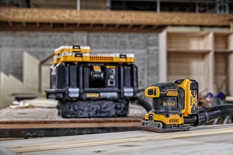 A DEWALT cordless sander (DCW200P2) is placed on a wooden plank, with a DEWALT tool storage box in the background inside a workshop setting. The sander is connected to a hose for dust extraction.