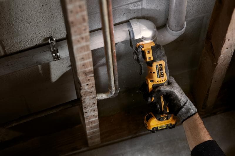Uma pessoa usando uma luva utiliza uma ferramenta multifuncional oscilante sem fio DEWALT para cortar um tubo de PVC branco perto de uma parede com tubos e vigas expostos.