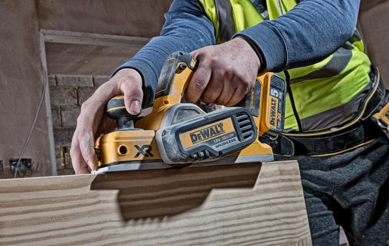 Uma pessoa está usando uma plaina sem fio DEWALT DCP580N para alisar a superfície de uma tábua de madeira. A ferramenta é amarela e preta, com marca e bateria visíveis.