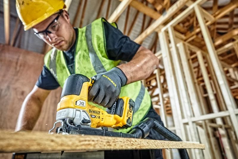 Uma pessoa usando capacete amarelo, colete de segurança e luvas está usando uma serra tico-tico sem fio DEWALT DCS334P2 para cortar uma tábua de madeira em um canteiro de obras com estrutura de madeira inacabada.