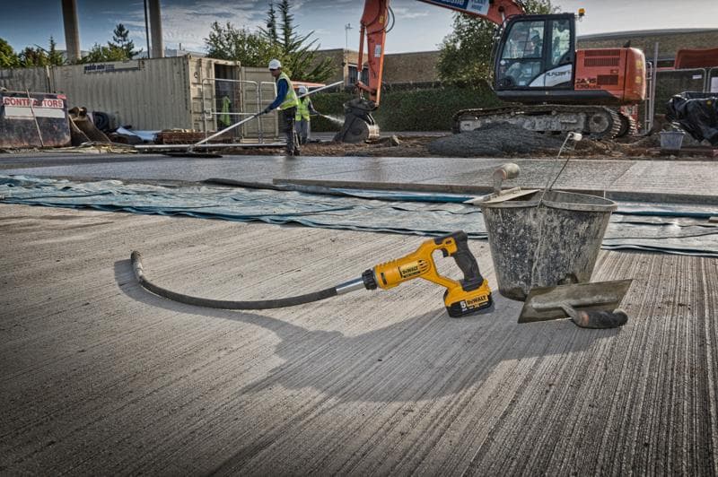 Uma ferramenta vibradora de concreto DEWALT DCE531N está colocada em um canteiro de obras, ao lado de um balde de metal e uma desempenadeira sobre uma superfície de concreto recém colocada. Ao fundo, há trabalhadores, uma escavadeira grande e outros materiais de construção.