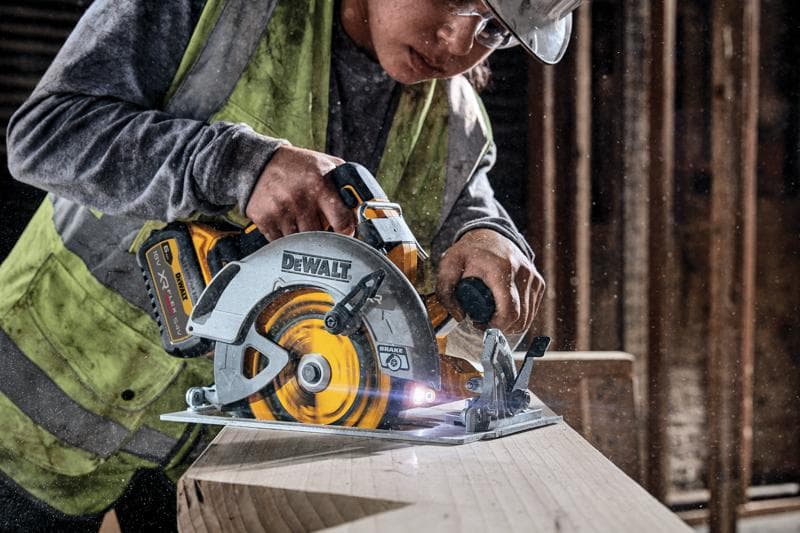 Uma pessoa usando colete de segurança e capacete está utilizando uma serra circular sem fio DEWALT DCS573NT para cortar uma tábua de madeira em um ambiente de construção.