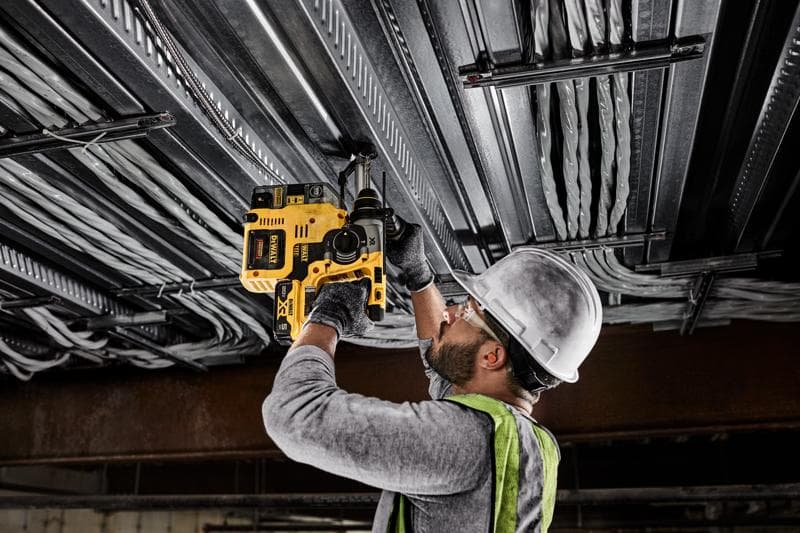 Uma pessoa usando capacete, óculos de proteção, luvas e colete de segurança utiliza um martelete rotativo amarelo DEWALT sem fio para perfurar um teto metálico com fiação elétrica.