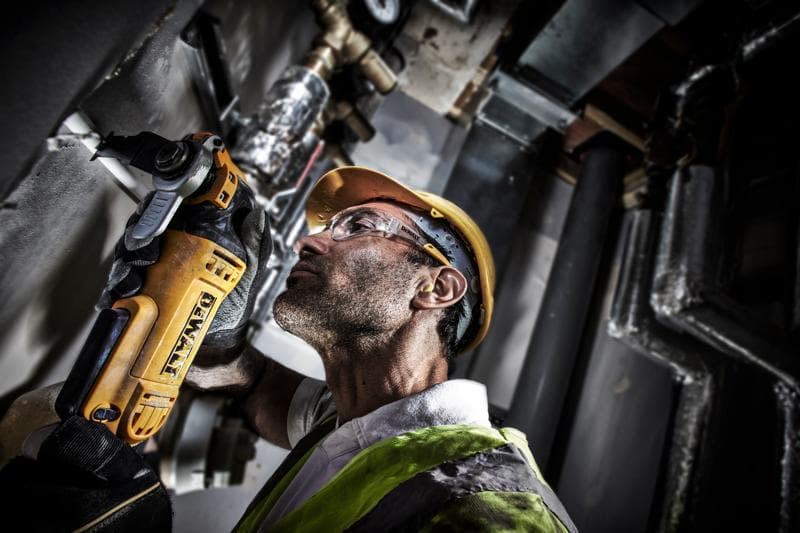 Uma pessoa usando capacete amarelo e colete de segurança utiliza uma ferramenta elétrica DEWALT DWE315 para cortar ou lixar material em uma parede em um ambiente industrial. O rosto está oculto.