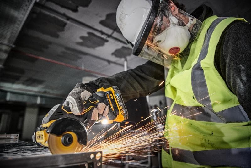 Uma pessoa usando equipamentos de segurança, incluindo capacete, viseira, máscara, luvas e colete de alta visibilidade, está usando uma ferramenta elétrica sem fio DEWALT DCS438N para cortar metal, gerando faíscas.
