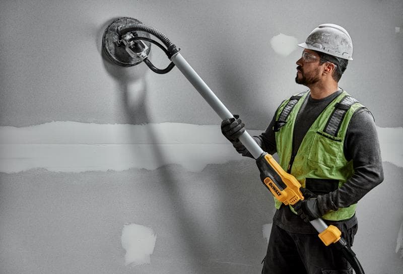 Um trabalhador da construção civil usando capacete e colete de segurança utiliza uma lixadeira de drywall DEWALT DWE7800 com cabo telescópico para lixar uma parede cinza.