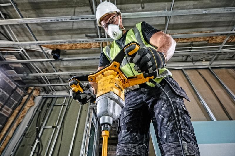 Um trabalhador da construção civil usando colete de segurança, luvas e capacete opera um misturador elétrico DEWALT DWD241 em um canteiro de obras, cercado por estruturas de metal e paredes inacabadas.
