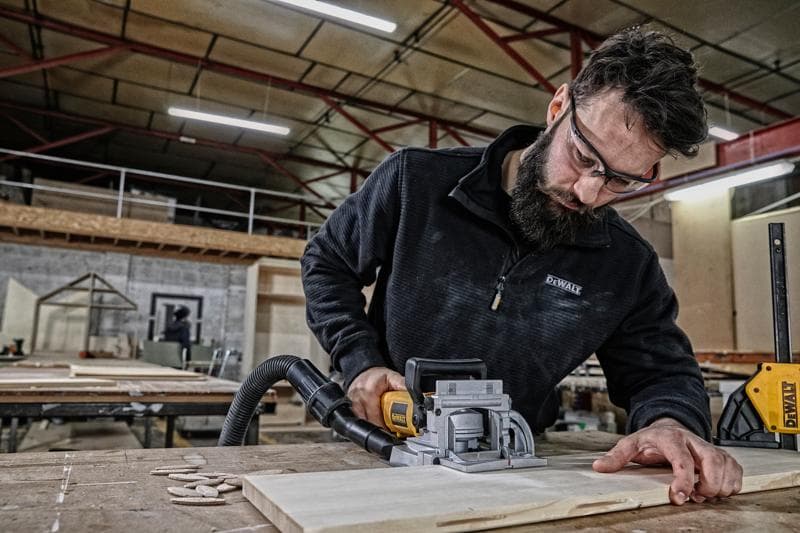 Uma pessoa usando uma ferramenta de marcenaria DEWALT DW682K para cortar uma peça de madeira sobre uma mesa em uma oficina. O rosto da pessoa está oculto para privacidade.