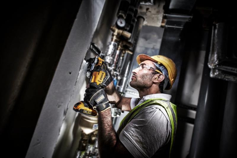 A construction worker wearing protective gloves, a hard hat, and a safety vest uses a DEWALT DCS355D2 cordless power tool to cut or grind a section of a wall or pipe in an industrial setting.