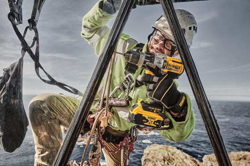 Um trabalhador usando equipamentos de segurança, incluindo luvas e capacete, utiliza uma furadeira sem fio DEWALT DCD996P2 em um local elevado ao ar livre perto do mar, com cordas e cinto de segurança visíveis.
