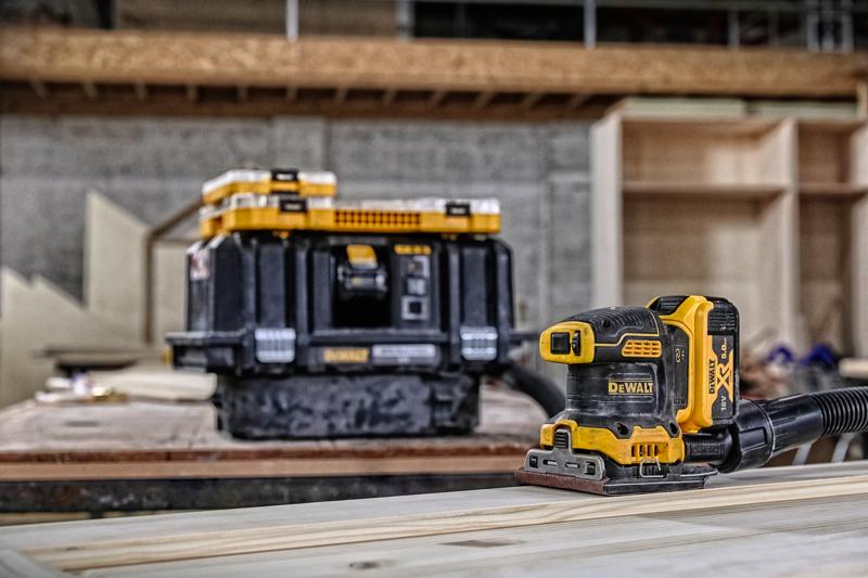 A DEWALT cordless sander (DCW200P2) is placed on a wooden surface, with a DEWALT tool storage box in the background. The