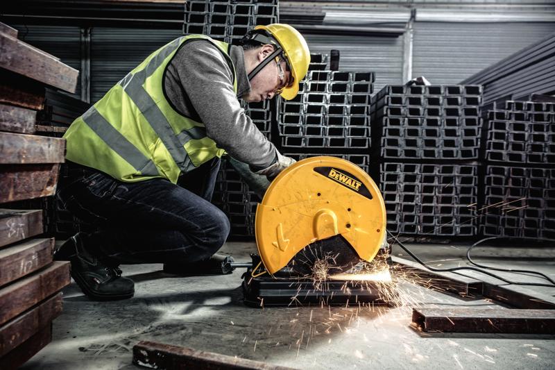 Uma pessoa usando equipamento de segurança opera uma serra de corte de metal DEWALT, modelo D28730, para cortar barras de metal em um ambiente industrial. Faíscas saem da área de corte.