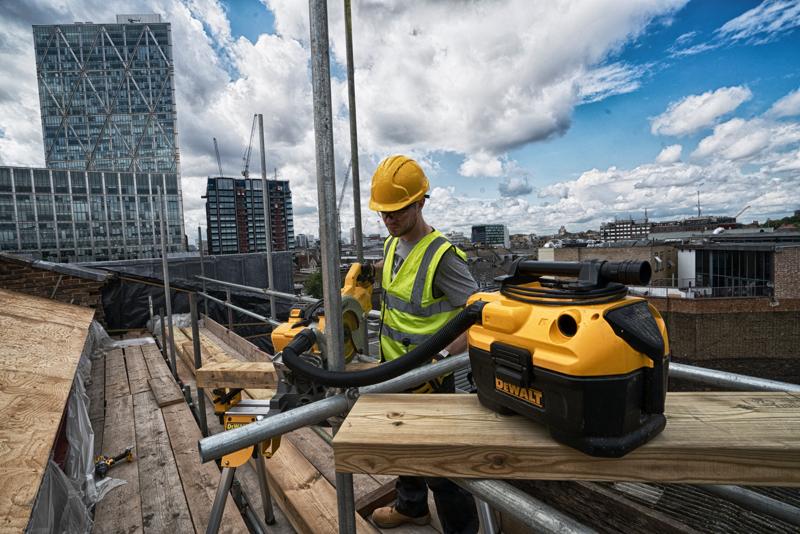 Um aspirador DEWALT DCV584 colocado em um canteiro de obras, com um trabalhador usando colete de alta visibilidade e capacete operando equipamentos ao fundo. Prédios altos e um céu parcialmente nublado são visíveis.
