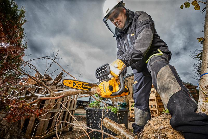 Uma pessoa com equipamento de segurança utiliza uma motosserra sem fio brushless DEWALT DCM565N para cortar galhos de árvores ao ar livre. A cena apresenta céu nublado, paletes de madeira e equipamento de proteção.