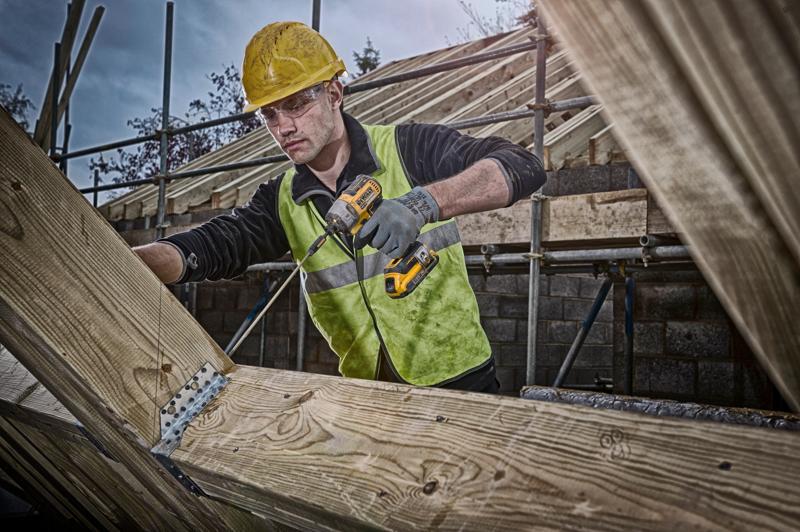 A construction worker wearing a yellow hard hat and reflective vest uses a DEWALT cordless drill to fasten metal brackets on wooden beams at a building site. His face is blurred.