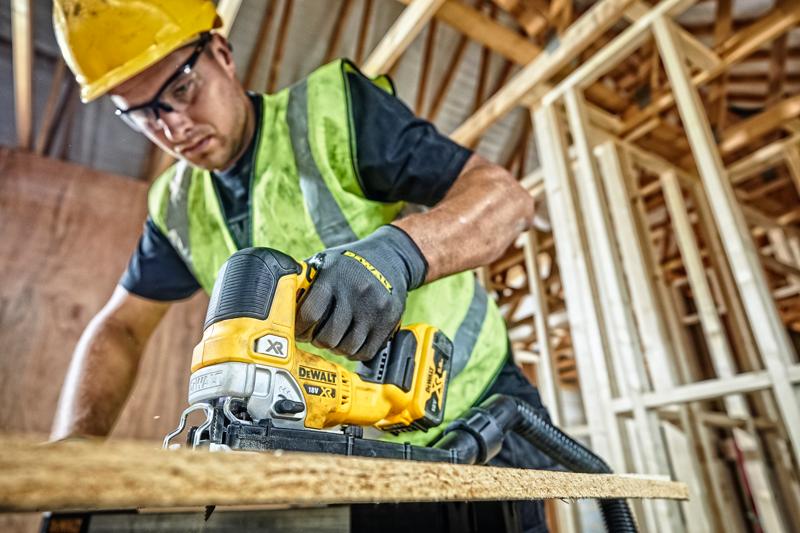 Uma pessoa usando capacete amarelo, colete de segurança e luvas está usando uma serra tico-tico sem fio DEWALT DCS334P2 para cortar uma tábua de madeira em um canteiro de obras com estrutura de madeira inacabada.