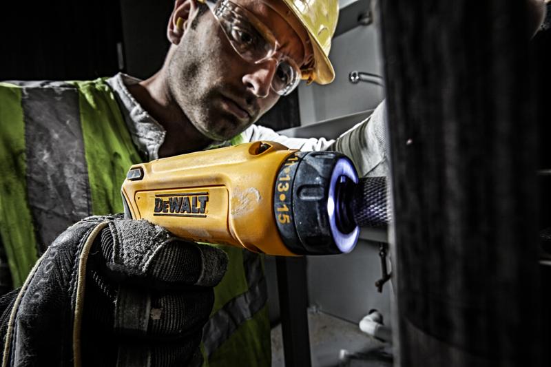 Uma pessoa usando luvas e colete de segurança utiliza uma chave de fenda elétrica amarela DEWALT (modelo DCF680G2) com ponta iluminada, trabalhando em um equipamento em ambiente industrial.