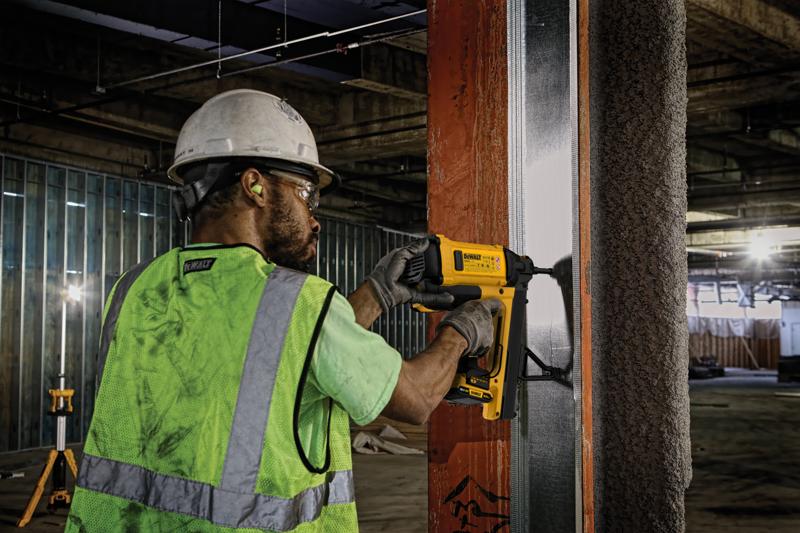 Um trabalhador da construção civil usando capacete de segurança, proteção auditiva, luvas e colete refletivo utiliza uma pistola de pregos DEWALT para fixar uma estrutura metálica em uma viga de aço em um canteiro de obras interno.