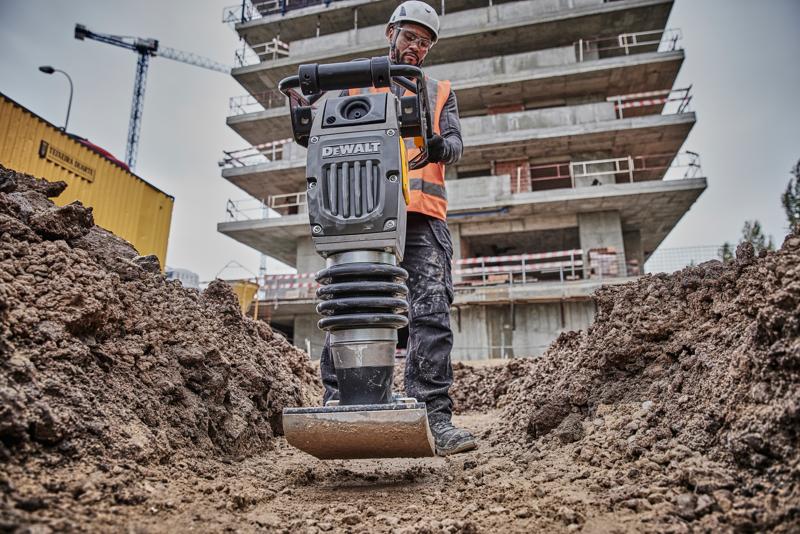 A person wearing a safety helmet and vest operates a DEWALT compactor tool in a dirt trench at a construction site, with a