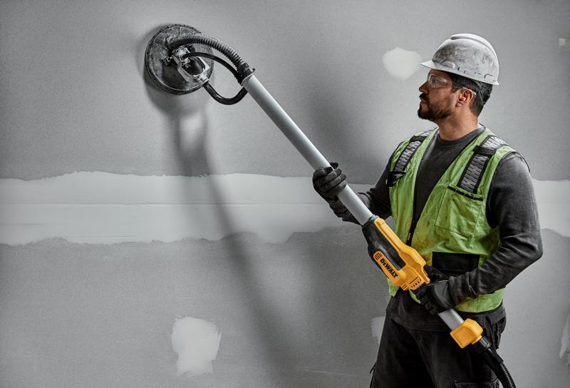 Um trabalhador da construção civil usando capacete e colete de segurança utiliza uma lixadeira de drywall DEWALT DWE7800 com cabo telescópico para lixar uma parede cinza.